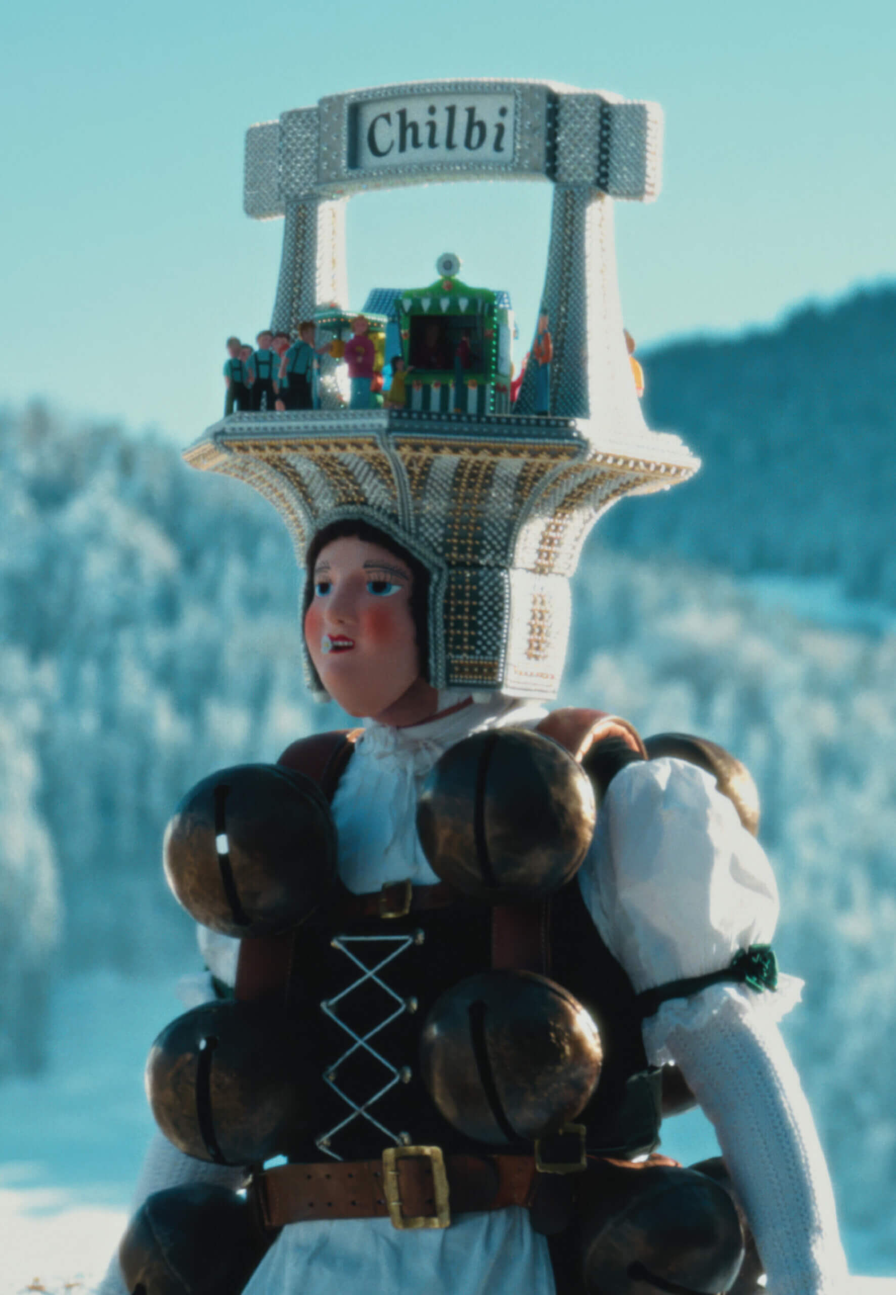 A portrait of a person wearing a human-faced mask, bells on front and back, and a diorama with a sign that reads “Chilbi.”