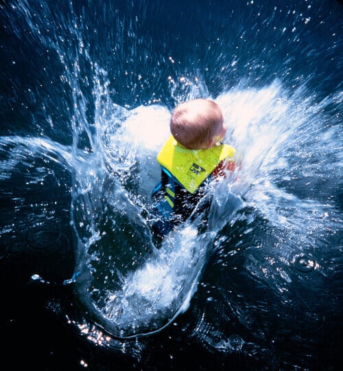 A photo depicts a blond baby wearing a bright yellow life jacket, splashing into a dark blue body of water.