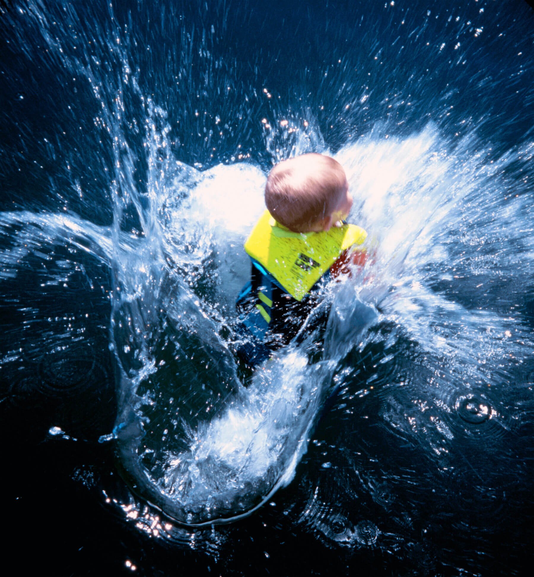 A photo depicts a blond baby wearing a bright yellow life jacket, splashing into a dark blue body of water.